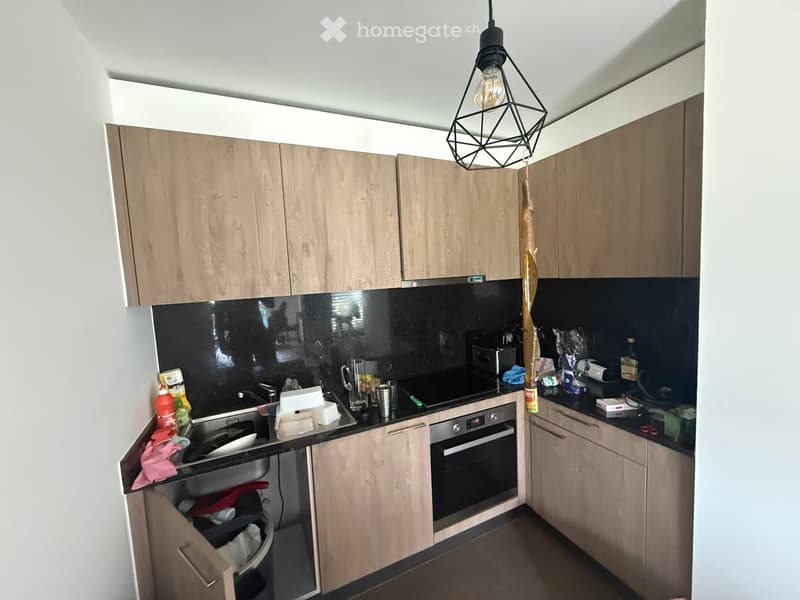 Modern kitchen with light wooden cabinets, black countertops, and stainless steel appliances. Visible items include a sink, oven, and various kitchen items on the counter.