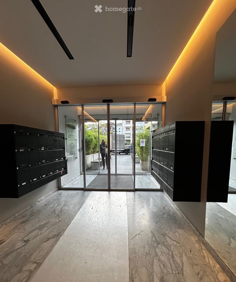 Modern hallway with polished marble floors, illuminated walls, and glass doors leading outside. Mailboxes are mounted on both sides of the corridor.