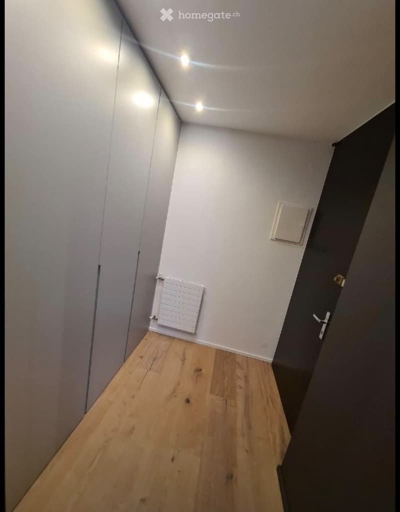 A narrow hallway with wooden flooring, white walls, and a radiator on one side. The hallway has a closed door at the end and a light fixture on the ceiling.