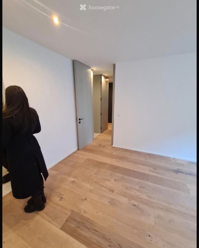Empty hallway with light wooden flooring and white walls, featuring an open doorway leading to another room.