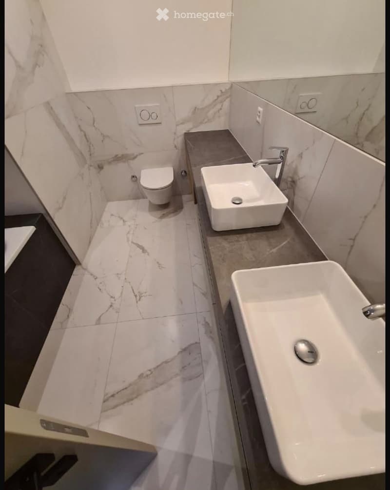 Modern bathroom with marble tiles, white toilet, two white sinks, and chrome faucets.