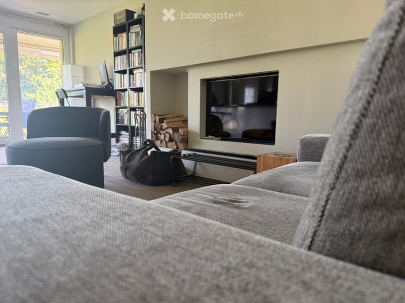 Modern living room with a grey sectional sofa, ottoman, armchair, coffee table, flat-screen TV, bookshelf, laptop, fireplace, and wood stack.