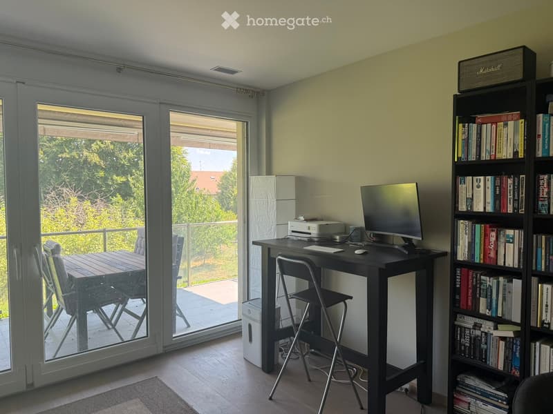 Bright room with a desk setup, a bookshelf, and a sliding glass door leading to a balcony.