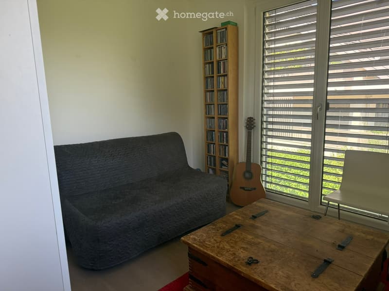 Living room with a dark grey sofa, wooden coffee table, guitar, bookcase, and a chair near the window with blinds.