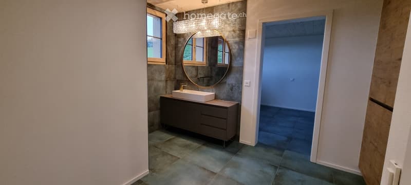 Modern bathroom with a dark grey cabinet, white sink, round mirror, and a chandelier above.