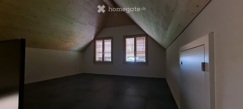 Empty attic room with wooden slanted ceilings, two windows with blinds, and a closed white door.