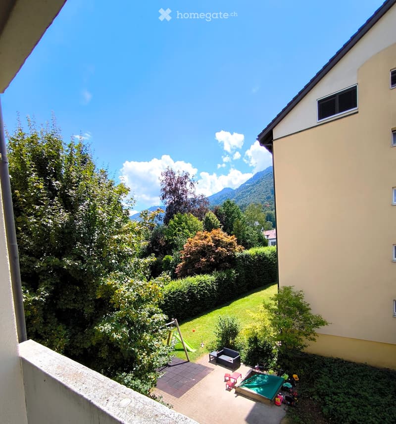 Balcony view overlooking a well-maintained garden with a small seating area, playground equipment, and a swimming pool, surrounded by trees and mountains.