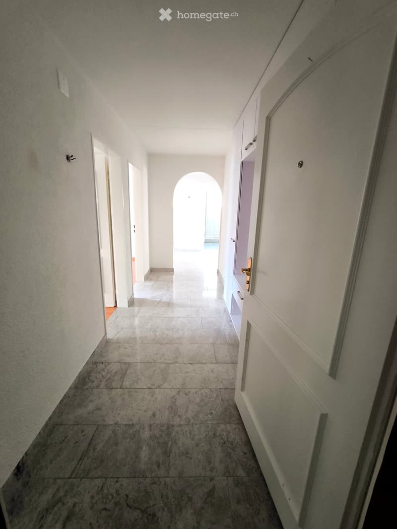 A long, narrow hallway with white walls, marble flooring, and several doors leading to other rooms.