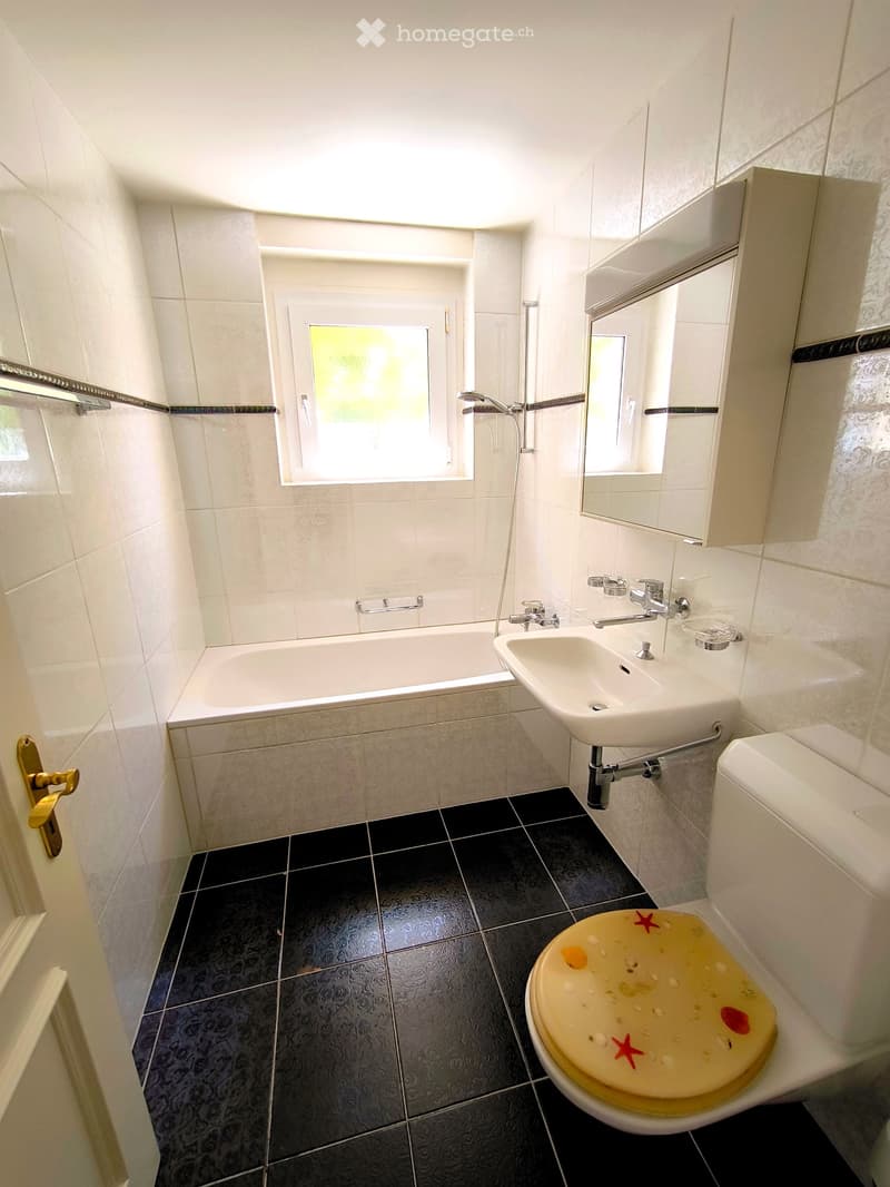 Modern bathroom with white tiles, black floor tiles, bathtub, sink, toilet, mirror, and cabinet.