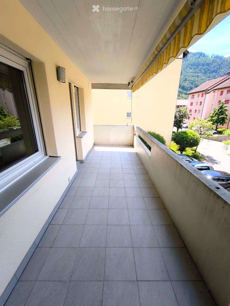 Balcony with tiled floor, beige walls, and a yellow awning. Offers a view of a green, hilly landscape with trees and parked cars.