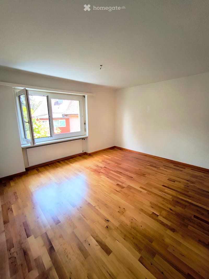 Empty room with wooden flooring, white walls, and a large window with white frame and radiator below it.