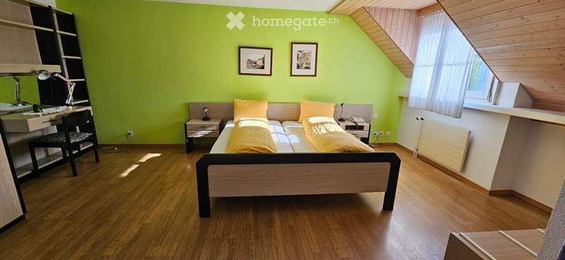Bright bedroom with wooden flooring, slanted wooden ceiling, and large window with white curtains. Features a double bed with yellow bedding, a nightstand, a desk with a chair, and a radiator.