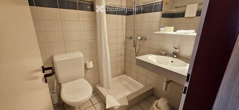 Clean, modern bathroom with white tiles, a toilet, a sink, a shower, a mirror, and a towel on the floor.