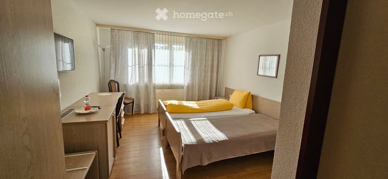 Cozy bedroom with two single beds, a desk with a laptop, a chair, and a television mounted on the wall. The room features wooden flooring and large windows with curtains.
