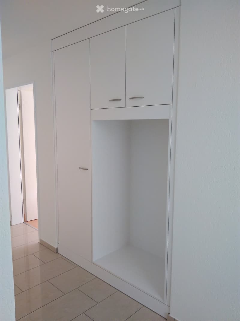 Empty closet with white built-in cabinets and a tiled floor, adjacent to a hallway with a visible doorway.