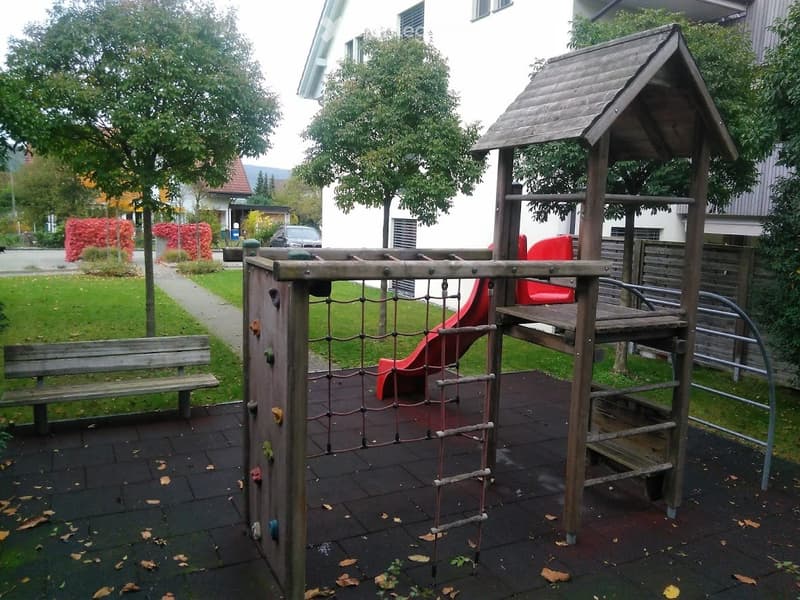 A small playground area with a climbing net, slide, and wooden playhouse, surrounded by a paved surface and greenery.