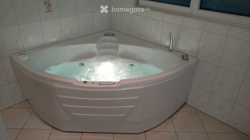 Modern corner bathtub with built-in jets and a sleek design, surrounded by tiled walls and a window above.