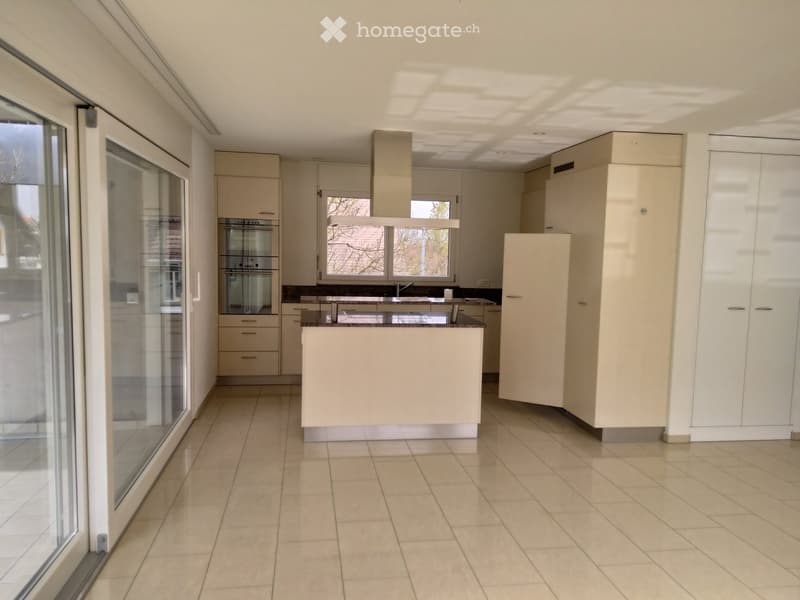 Modern kitchen with light-colored cabinets, a central island, and a built-in oven. The space features tiled flooring and large windows allowing natural light.