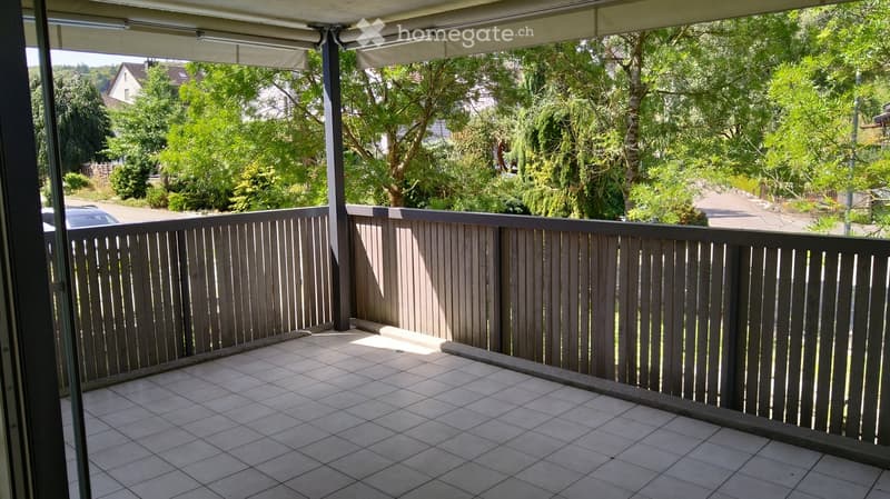 Balcony with tiled floor, wooden railing, and retractable awning, overlooking a green, tree-filled area.