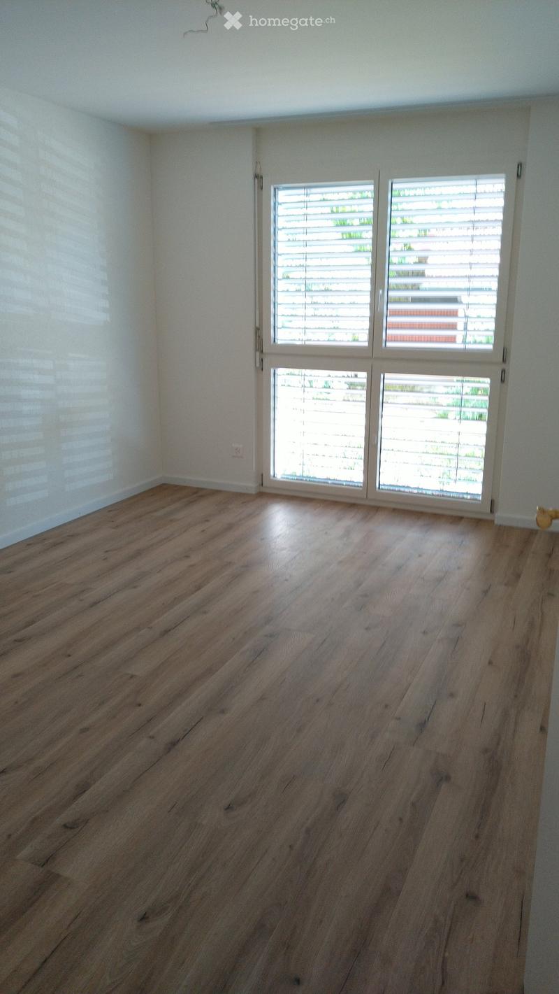 Empty room with wooden flooring and large windows with blinds, allowing natural light to enter.