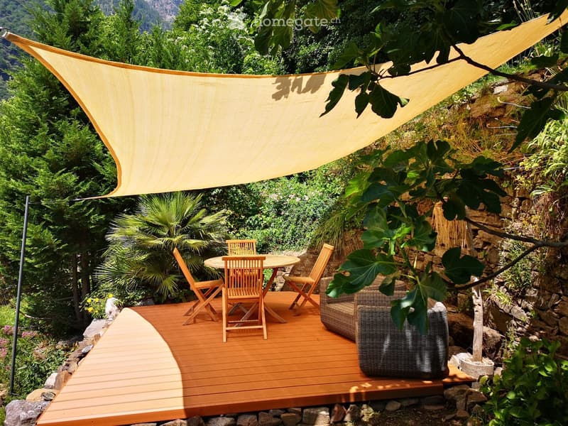 Outdoor wooden deck with table and chairs under a shade sail, surrounded by greenery and a stone wall