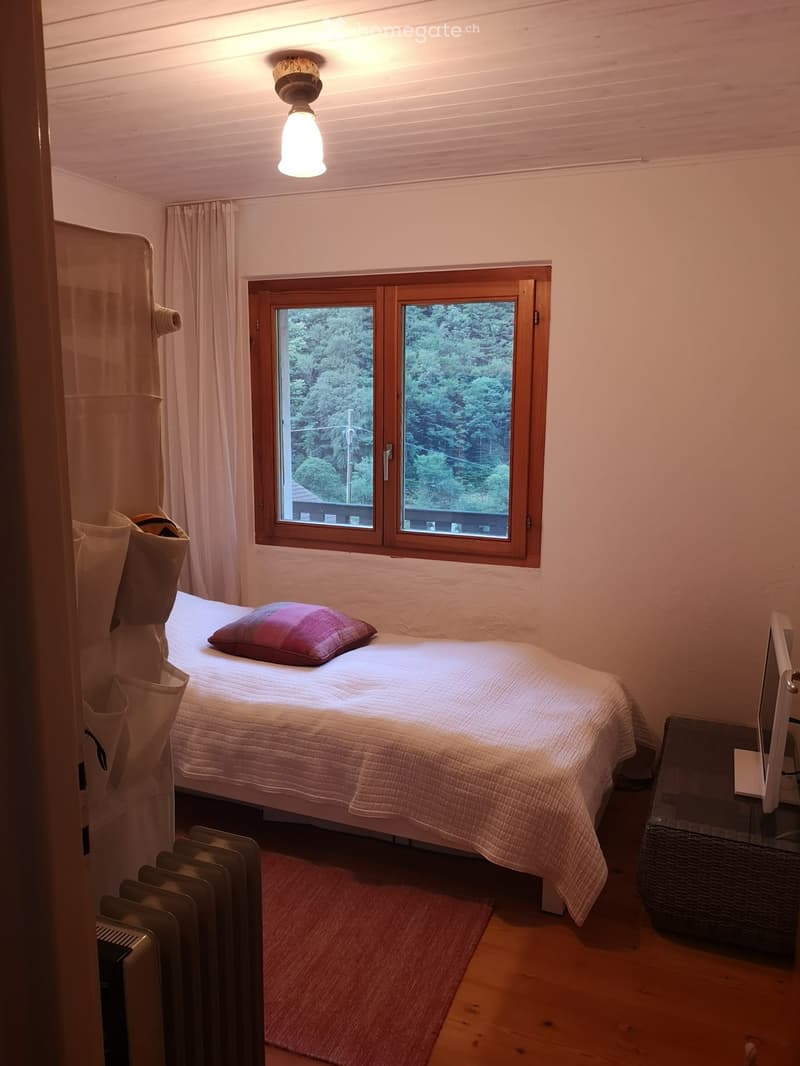 Single bed with a white quilt, a red pillow, a wooden window with curtains, a wooden side table, a monitor on top of the side table, a radiator, a rug, a wooden floor, a wooden ceiling with ceiling light.