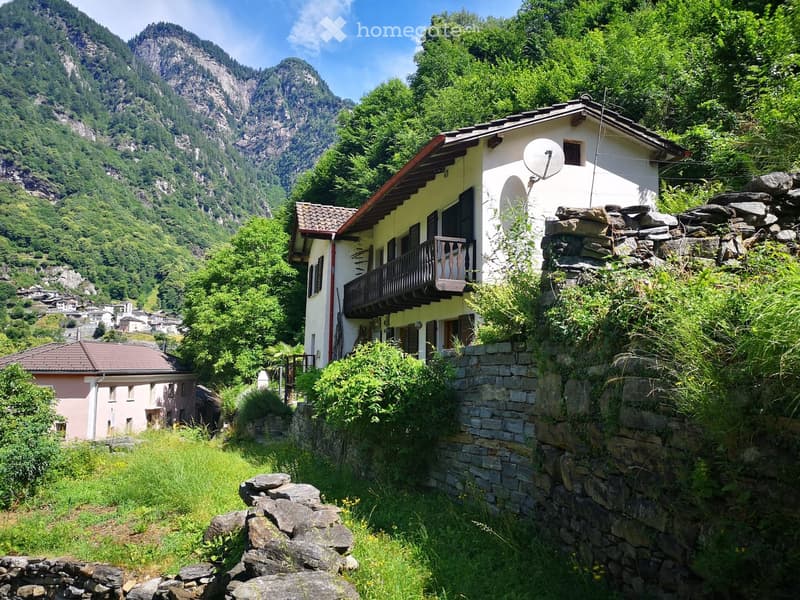 House located on a hillside, white exterior, red tile roof, balcony, mountains in the background, fenced by stone walls