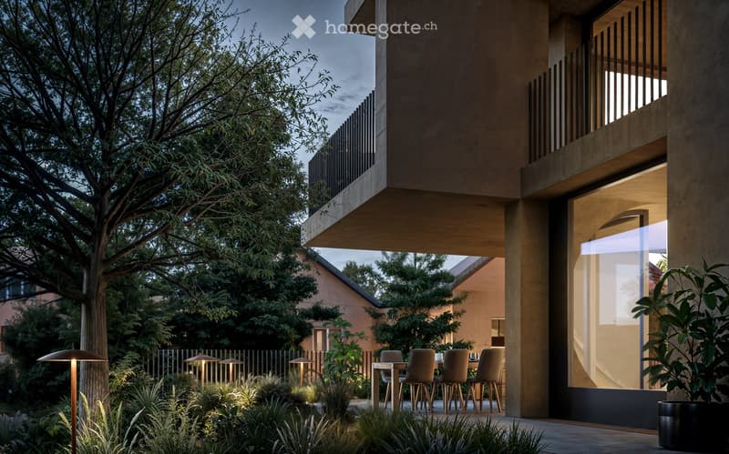 Covered outdoor patio area with wooden furniture, surrounded by lush greenery and trees. The patio is connected to the building through a glass wall, allowing natural light to enter the space.