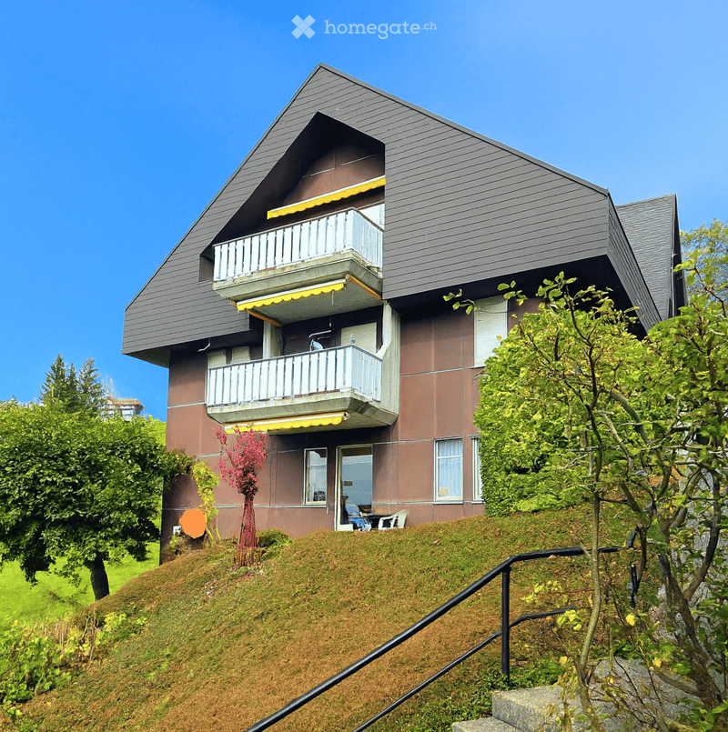 Two-story house with grey roof, brown facade, and multiple balconies with yellow awnings. The house is situated on a raised ground floor with a staircase leading up to it, surrounded by greenery and trees.