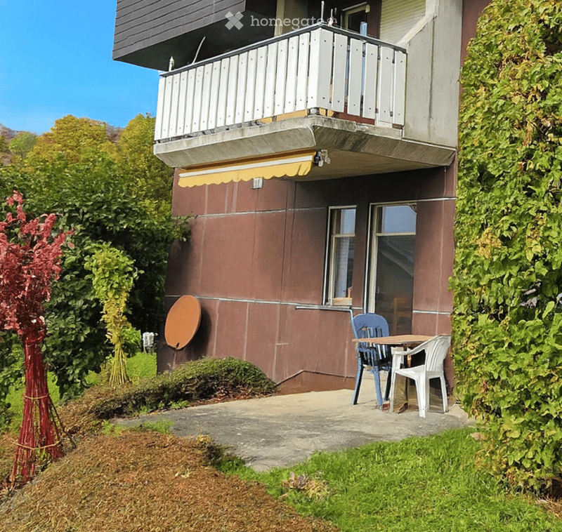 Balcony with a small seating area, including a table and two chairs, surrounded by greenery and a clear blue sky.