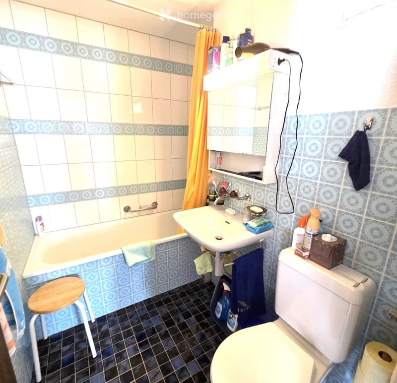 Bathroom with a bathtub, sink, toilet, mirror, and a small wooden stool. The walls are tiled in blue and white, and there are towels and cleaning supplies visible.