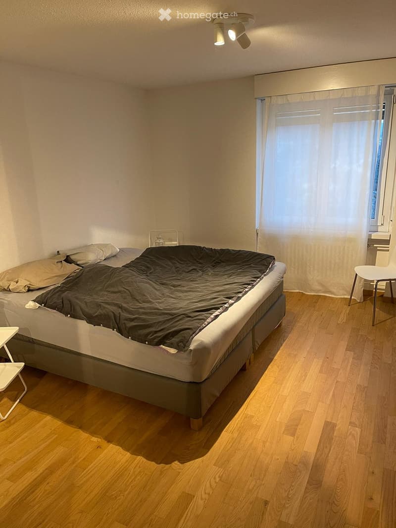Minimalist bedroom with a single bed, two small stools, and a window with sheer curtains. Light wooden flooring and white walls.
