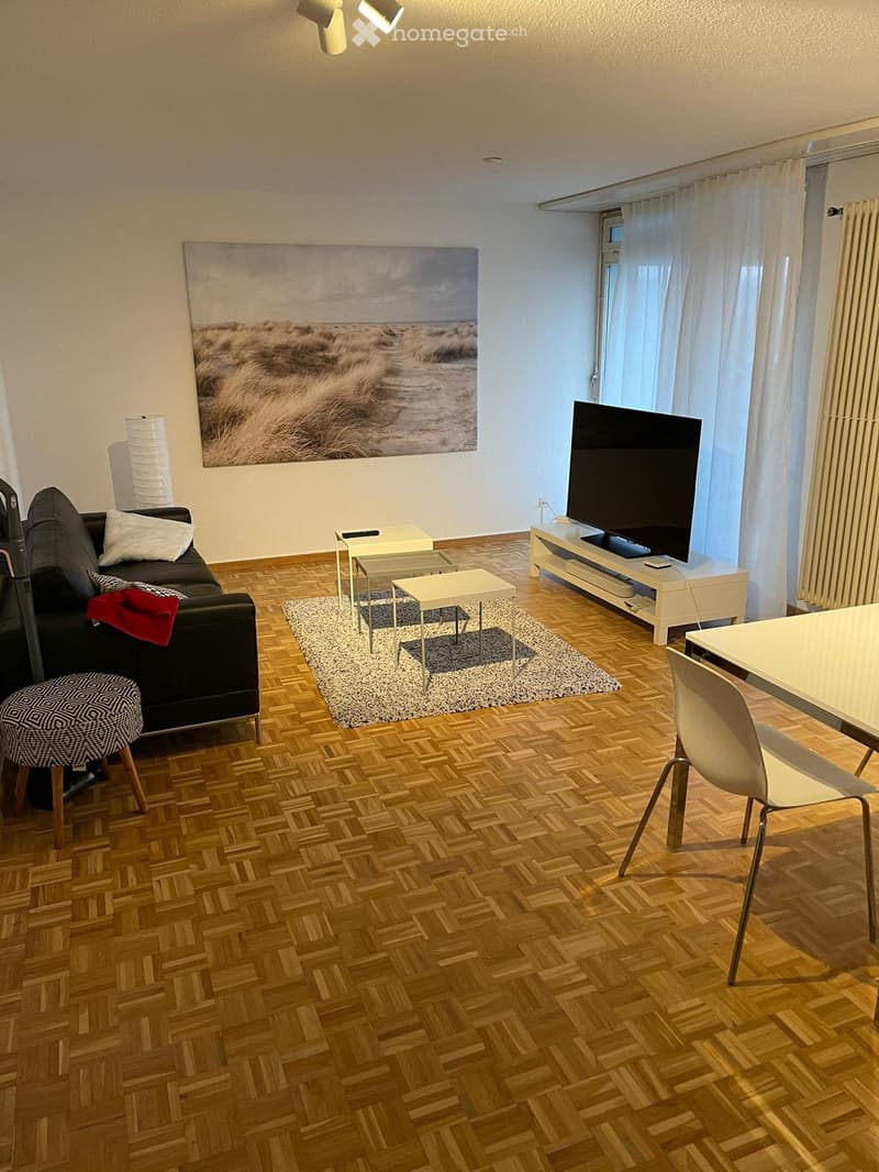 Modern living room with wooden herringbone flooring, a black sofa with pillows, a patterned stool, a coffee table, a TV on a stand, and a dining area with a table and chairs.