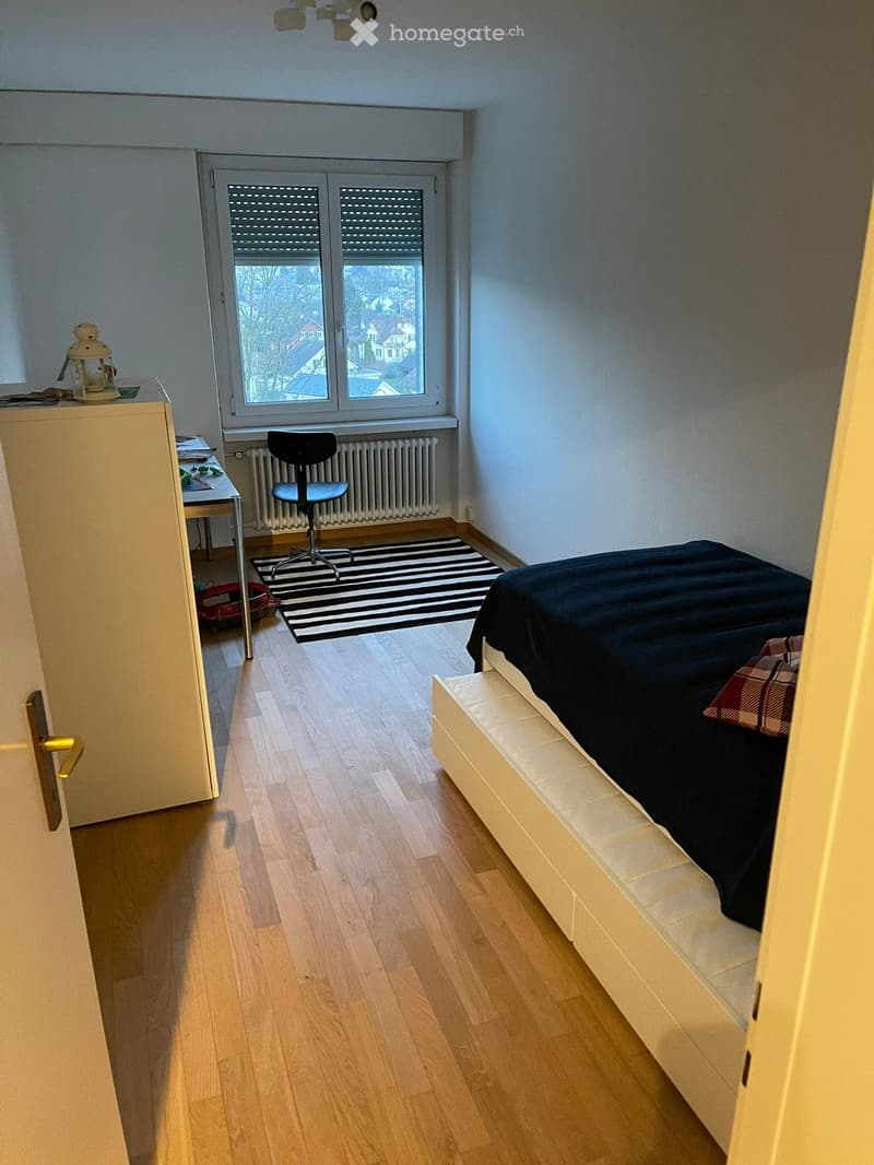 A well-lit bedroom with wooden flooring, a bed with dark bedding, a chair, a small table with a lamp, and a radiator under the window.