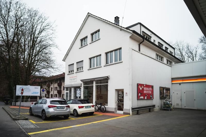 A two-story white building with a pitched roof, multiple windows, and a storefront with glass doors. There are two parked cars in front, a bicycle, and a bench near the entrance.