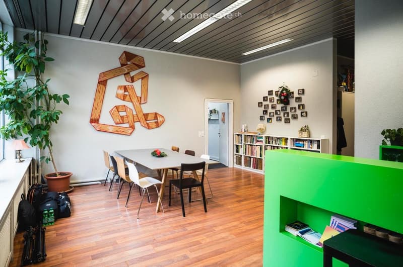 Bright, modern room with wooden flooring, a large table surrounded by chairs, a bookshelf filled with books, a potted plant, and a decorative wall piece.