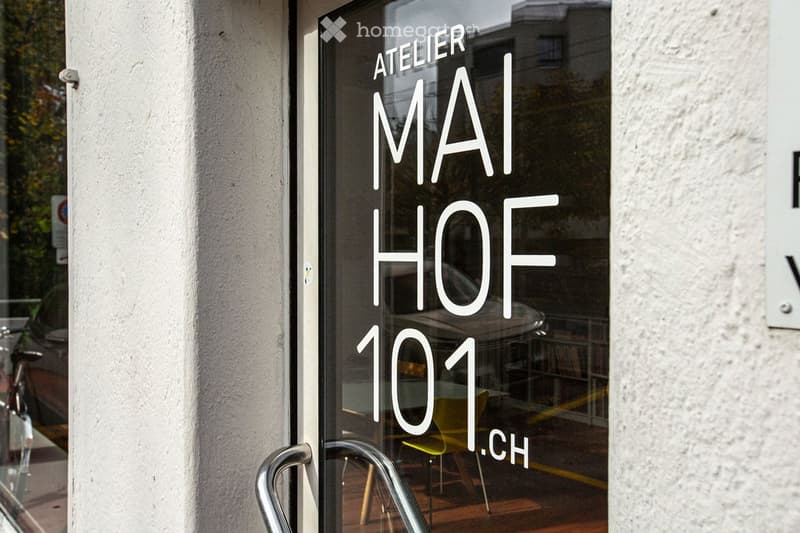 Storefront with glass door labeled "ATELIER MAI HOF101.CH" and a reflection of interior furniture visible through the glass.