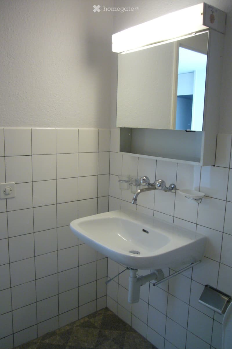 A small bathroom with a white sink, faucet, mirror, and two cups on the sink counter. The walls are tiled with white tiles, and there is a light fixture above the mirror.