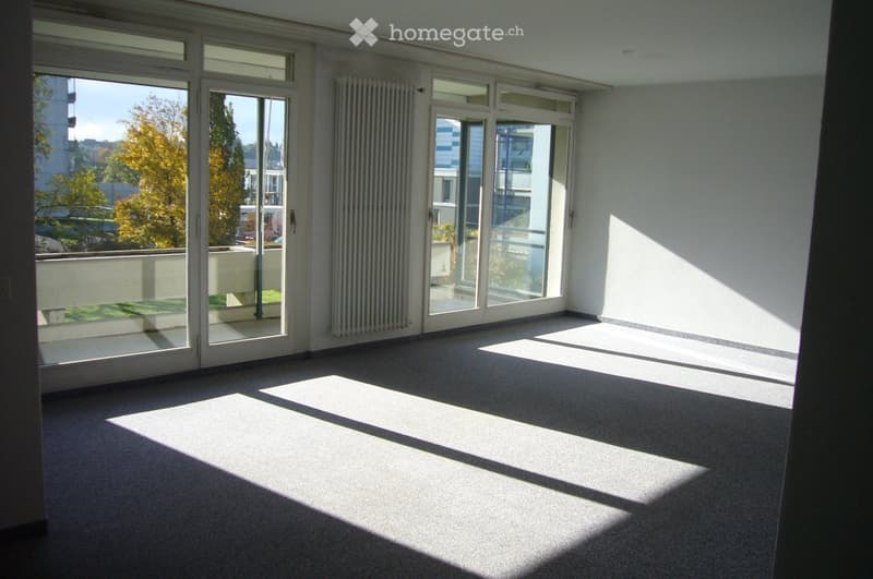 Empty room with large windows and sliding glass doors leading to a balcony, carpeted floor, radiator, and natural light streaming in.