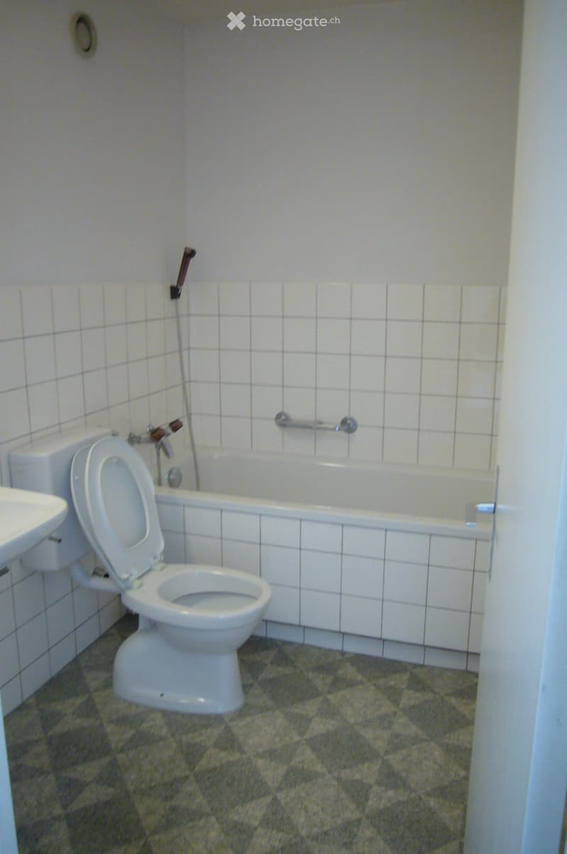 Bathroom with white tiled walls, a toilet, a bathtub, a sink, and a faucet. The floor is covered with a patterned tile.