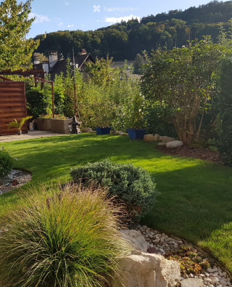 Well-maintained garden with a lawn, various plants, shrubs, and a wooden fence. Features include a lantern, potted plants, and a stone pathway.