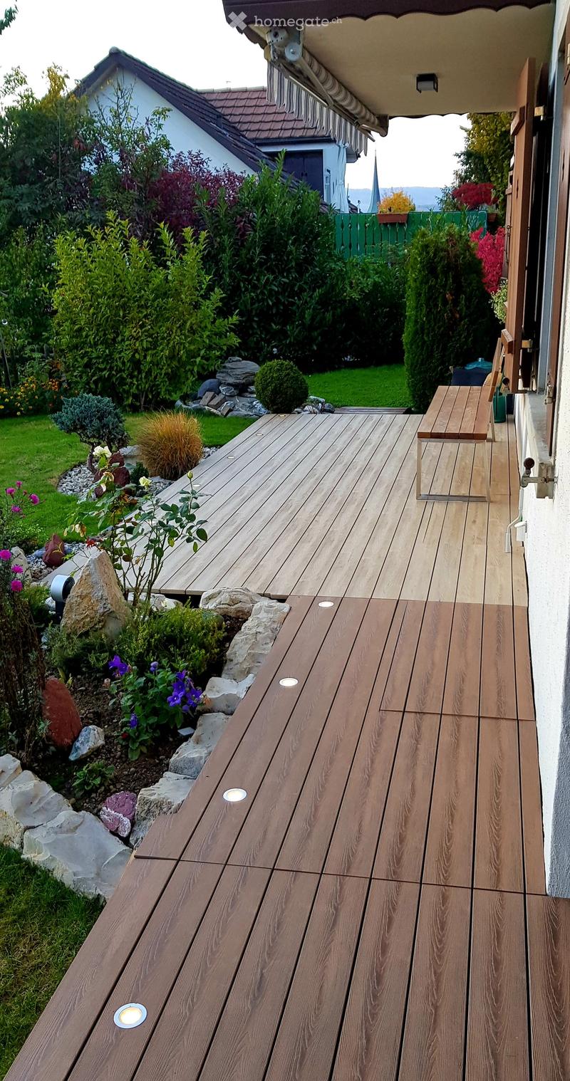 Wooden deck with built-in LED lights, small wooden bench, surrounded by garden with various plants and shrubs.