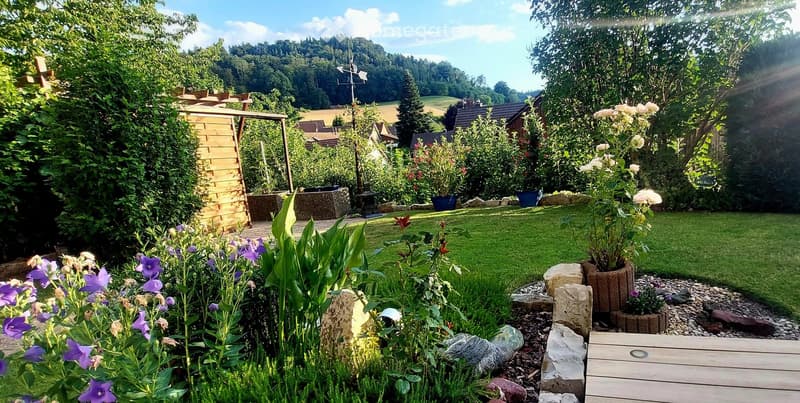 Well-maintained garden with a variety of flowers, shrubs, and trees, featuring a wooden deck, stone elements, and a weather station in the background.