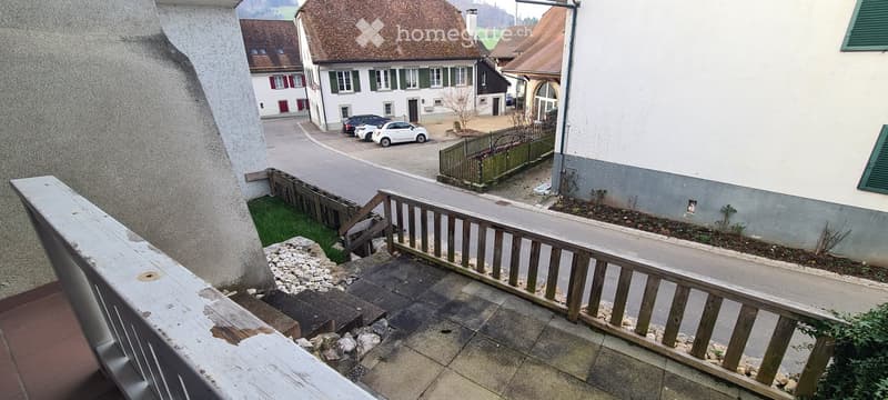 concrete balcony with wooden railing and steps leading to terrace with paved ground, fenced parking lot, view of street with cars and buildings