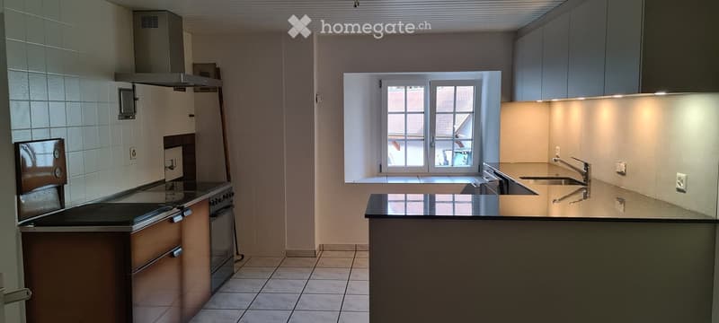 Modern kitchen with granite countertop, gas stove, electric oven, sink with faucet, wooden cabinets, tiled floor, window, tiled backsplash, exhaust hood.