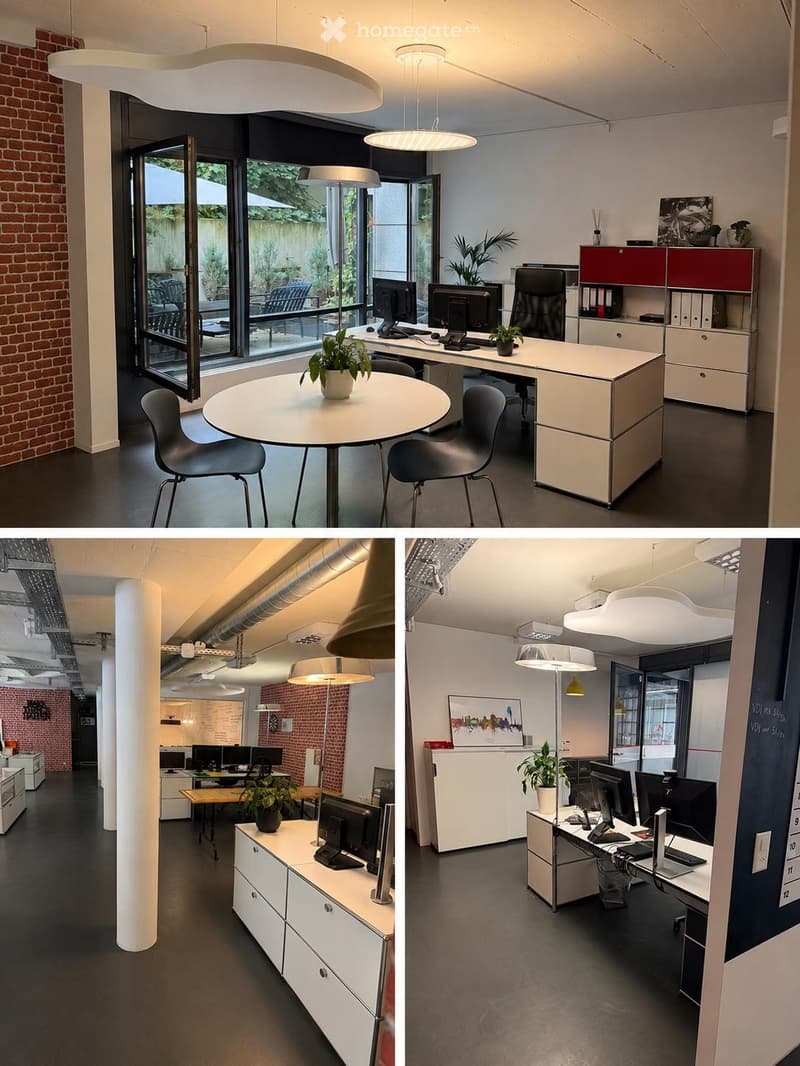 Modern office space with large windows, round meeting table, chairs, desks with computers, cabinets, and plants.