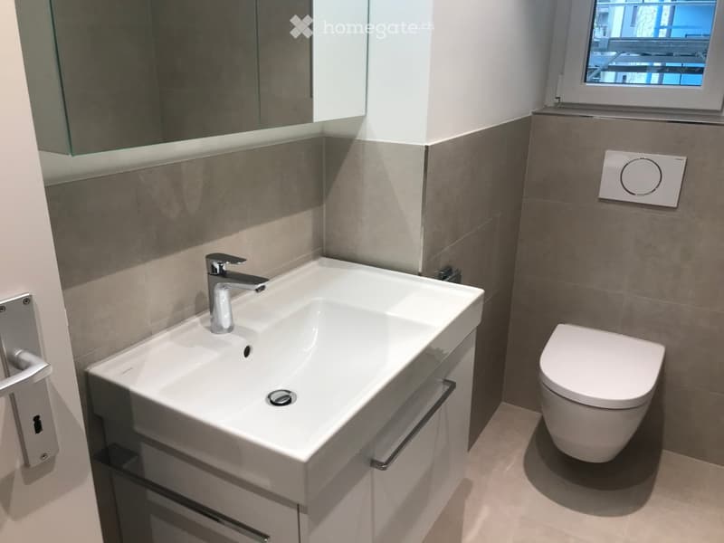 Modern bathroom with a white sink, chrome faucet, and toilet. Features a large mirror and a window with a view.