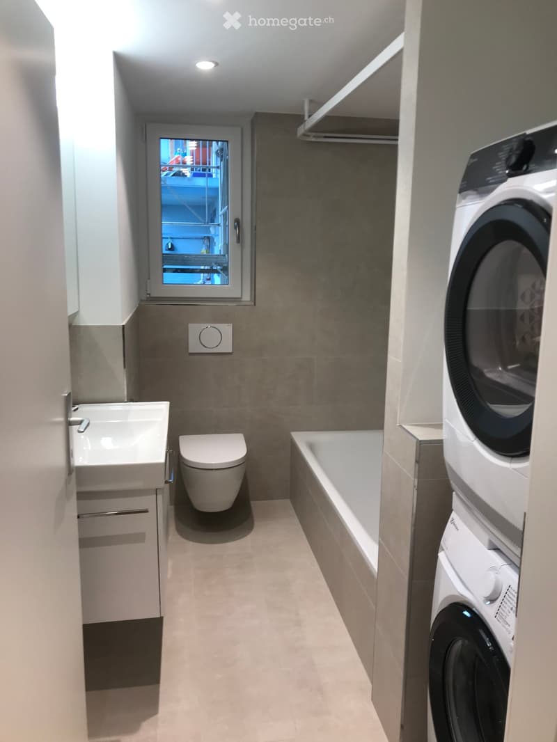 Modern bathroom with a sink, toilet, bathtub, and stacked washing machine/dryer unit. Features a window with a view of another room.