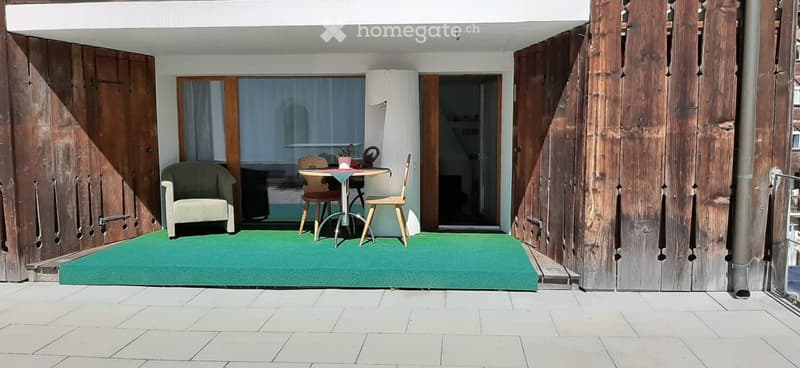 Balcony with green artificial turf, wooden shutters, small table with chairs, and a view of the interior space.