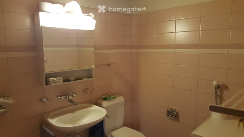 Bathroom with beige tiled walls, a sink, toilet, mirror, and a light fixture above the mirror. A towel is hanging on the side.